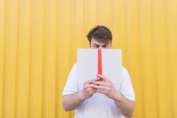 Смешной человек стоит на фоне цветной стены и закрывает лицо книгой. Портрет человека с книгой на желтом фоне . — стоковое фото