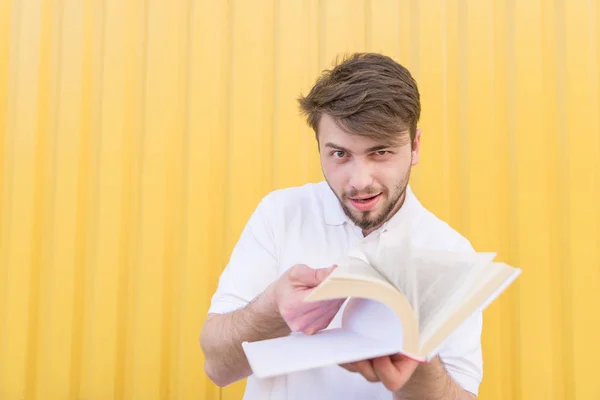 Забавный человек пролистывает страницы книги и смотрит на камеру на желтом фоне. Весёлый студент с книгой на фоне жёлтой стены — стоковое фото