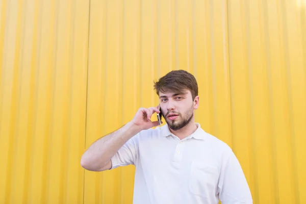 Um homem bonito fica no fundo de uma parede colorida, fala ao telefone e olha para a câmera. Portert de um marido sério durante um telefonema em um fundo amarelo — Fotografia de Stock