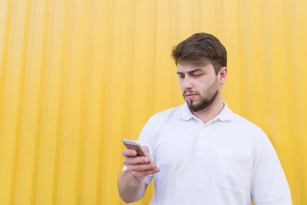 Homem adulto com barba fica no fundo de uma parede amarela e usa um smartphone — Fotografia de Stock