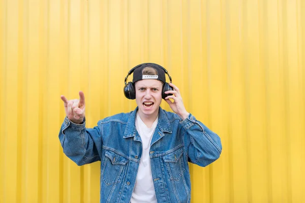 Um jovem com uma jaqueta de ganga está ouvindo rock em seus fones de ouvido e mostra um sinal de heavy metal. No fundo da parede amarela. Conceito musical — Fotografia de Stock