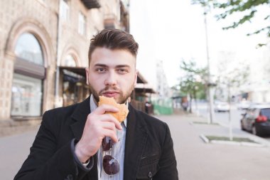 Sokak bir arka plan elinde sandviç ile elbiseli sakallı bir adam portresi. Meşgul bir adam caddede gidiyor ve kahvaltı için atıştıracak bir şeyler yiyor.