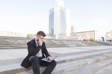 Kentsel manzara fonunda dizlerinin üzerinde bir dizüstü bilgisayar ile bir merdiven üzerinde oturan düşünceli bir iş adamı. Sokakta bir bilgisayar olarak çalışır. İş kavramı. Son Tarih