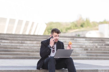 Meşgul bir adam iş takım elbiseli bir dizüstü bilgisayar ile dizüstü bilgisayarlarda kucağına oturur, bir sandviç yiyor ve bir cep telefonu konuşuyor. Bir işadamı çalışma ve sokakta hızlı yemek yeme.
