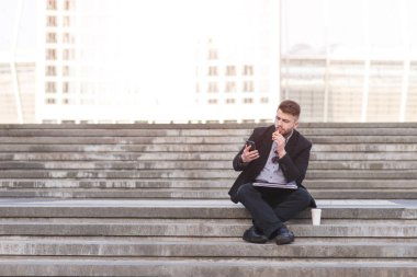 Düşünceli bir işadamı bir merdiven bir aperatif, bir fincan kahve ve bir balığı oturur. Bir iş adamı açık havada oturur, bir sandviç yiyor ve bir akıllı telefon kullanır