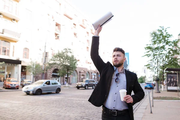 Деловой человек с документами и чашкой кофе в руках ловит такси у дороги. Мужчина опаздывает на работу, поэтому он ловит такси. — стоковое фото