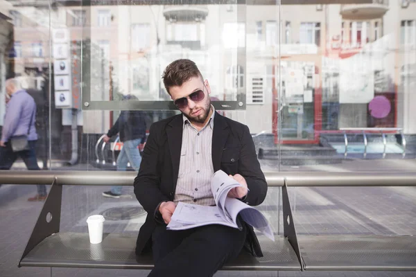 Un oficinista en un traje sentado en una parada de autobús y leyendo documentos. Un hombre de negocios con estilo en sus gafas y documentos en sus manos se sienta en el banco — Foto de Stock
