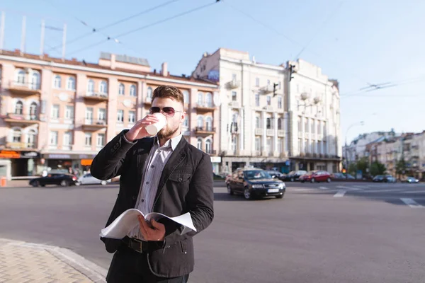 Бизнесмен стоит на фоне города с документами в руках и пьет кофе. Деловой человек пьет кофе из чашки на фоне городского пейзажа . — стоковое фото