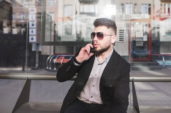 Бизнесмен сидит на автобусной остановке и разговаривает по телефону. Стильный мужчина в костюме говорит по телефону в общественном транспорте останавливает скамейку . — стоковое фото