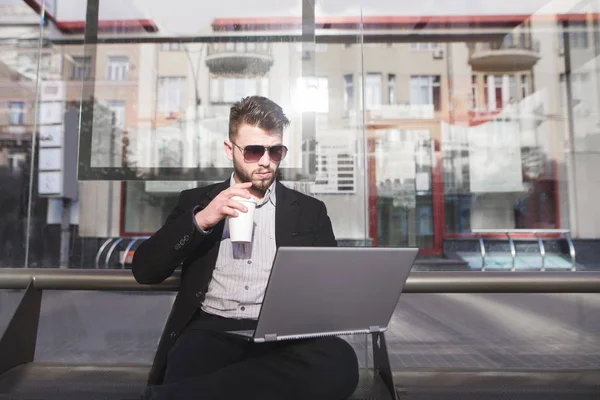 Зайнятий чоловік сидить за ноутбуком на зупинці і п'є каву. Ділова людина працює в громадському місці. Зайнятий чоловік не має часу на роботу. . — стокове фото