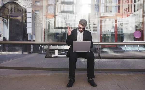 A busy man sits on a public transit stop and works on a laptop. Work on the way to the office. Businessman does not have time to do the job. Deadline concept.