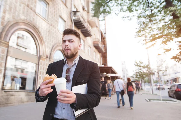 A busy man eats and drinks coffee while walking for work. Business man has breakfast with fast food. A businessman goes to work on the street and eats a sandwich