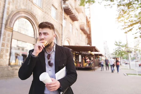 Um homem de negócios com documentos nas mãos come um lanche enquanto caminha para o trabalho. Um homem ocupado morde um sanduíche, bebe café e vai trabalhar . — Fotografia de Stock
