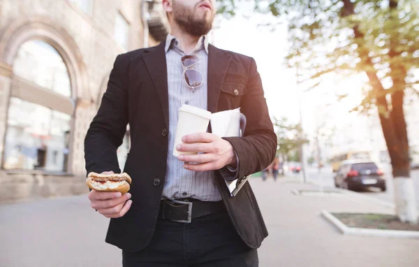 Бизнесмен идет по улице с закуской и кофе в руках. Занятый человек ест сэндвич по дороге на работу. Завтрак. Концепция бизнеса . — стоковое фото