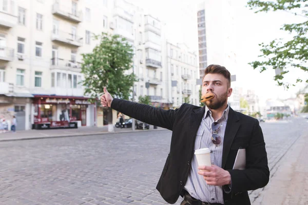 Um homem ocupado com uma xícara de café em suas mãos e uma sanduíche em seus dentes pega um táxi na estrada. Homem de negócios está com pressa e pequeno-almoço na estrada — Fotografia de Stock