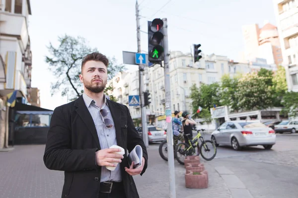 Bonito empresário com uma xícara de café e documentos em suas mãos está na travessia de pedestres e aguarda a luz verde. Um homem de negócios anda pela cidade — Fotografia de Stock