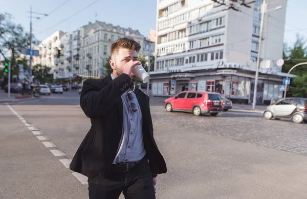 El hombre de negocios cruza la calle en un cruce peatonal y bebe café. Un hombre de negocios ocupado bebe café para desayunar de camino al trabajo . — Foto de Stock