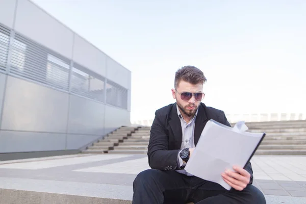Концентрований бізнесмен сидить на сходах і дивиться на папери в руках. Бородатий бізнесмен в окулярах читає документи на тлі міського пейзажу . — стокове фото