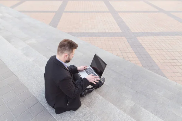 Un hombre de negocios está sentado en las escaleras y trabajando en un portátil. Un hombre de negocios ocupado trabaja al aire libre. Concepto de negocio. Fecha límite —  Fotos de Stock