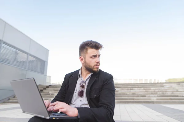 Affärsman sitter på en trappa med en laptop i knäet-fältet och verk. Den förbjudna man arbetar utomhus och ser bort. Affärsidé. — Stockfoto