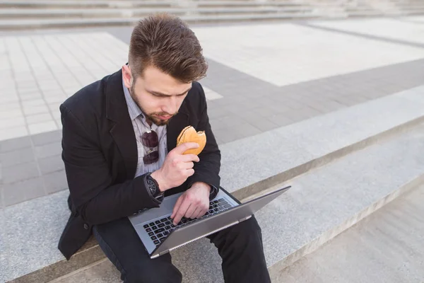 Зайнятий бізнесмен працює на блокноті і їсть бутерброд на відкритому повітрі. Зайнятий чоловік працює на ноутбуці і їсть закуску на вулиці . — стокове фото