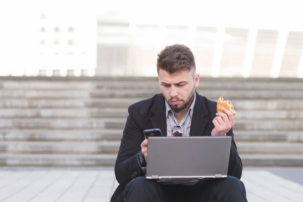 Зайнятий чоловік сидить на сходах з ноутбуком і закускою і використовує смартфон. Зайнятий бізнесмен працює на ноутбуці і їсть на відкритому повітрі . — стокове фото