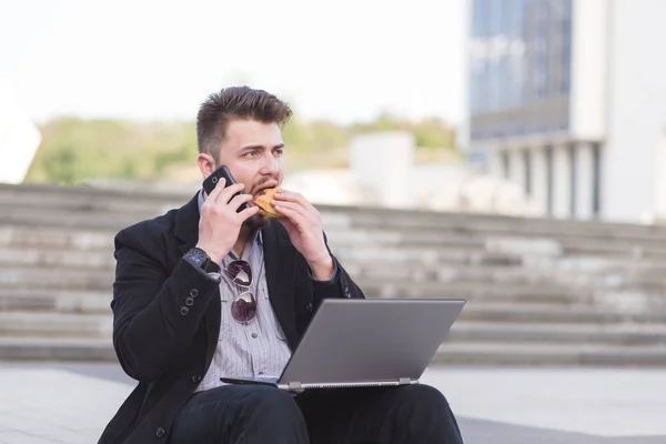 Businessman is sitting on a laptop-lap tops, eating a sandwich and talking on a mobile phone. A busy man works and snacks in the street. Business concept. — Stock Photo, Image