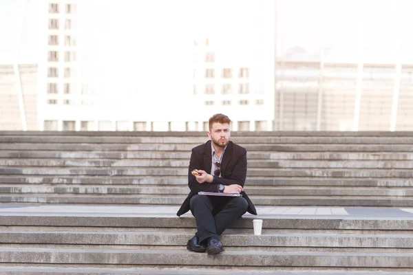 Busy man is sitting on the street with a snack and a cup of coffee in the background of the city landscape. Businessman sitting on the stairs and eating.