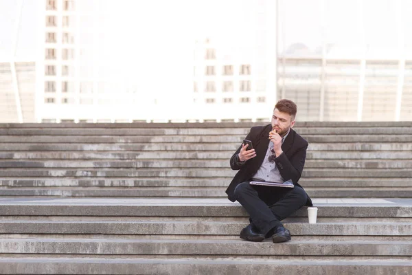 Задумчивый бизнесмен сидит на лестнице с закуской, чашкой кофе и снэппером. Бизнесмен сидит на открытом воздухе, ест сэндвич и пользуется смартфоном. — стоковое фото