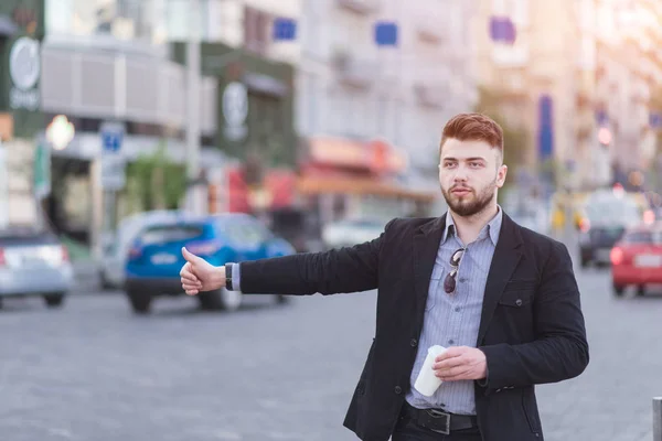 Egy szilárd szakállas férfi, egy öltöny áll az úton, egy csésze kávéval a kezében, és fogás egy taxit. Elfoglalt üzletember fogások az autó az úton — Stock Fotó