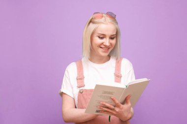 Elinde mor bir arka planda kitabı olan mutlu sarışın kız gülümseyip okuyor. Şirin elbiseli mutlu genç kız kitap okuyor ve gülüyor, pembe üzerinde izole edilmiş..