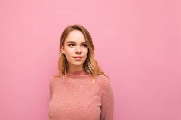 Hermosa chica rubia en un suéter rosa aislado sobre un fondo rosa, mira hacia un espacio en blanco con una sonrisa. Señora positiva mirando hacia otro lado en el fondo de color. Espacio de copia — Foto de Stock