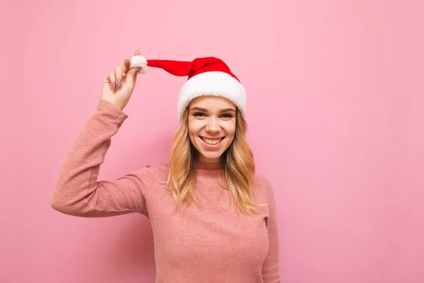 Улыбающаяся девушка в розовом свитере и шляпе Санта Клауса стоит на розовом фоне, смотрит в камеру и улыбается. Портрет счастливой блондинки в Рождество, изолированные на розовом фоне . — стоковое фото