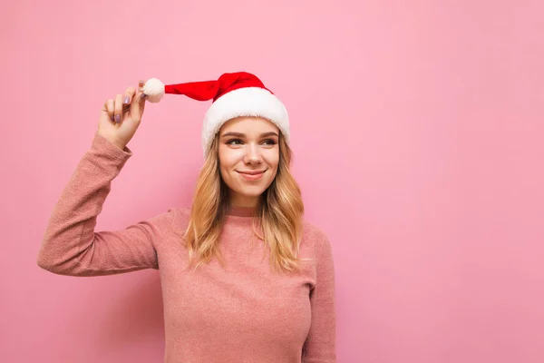Позитивная девушка в шляпе Санты стоит на розовом фоне, смотрит в пустоту и улыбается. Счастливая блондинка в Рождество изолирована на розовый. Концепция Рождества — стоковое фото