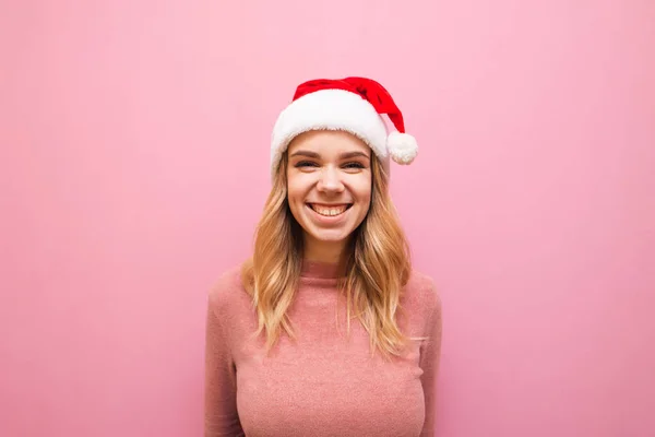 Портрет улыбающейся девушки в рождественской шляпе на розовом фоне, смотрит в камеру и смеется. Радостная леди Санта улыбается, одетая в розовый теплый свитер и шляпу Санты. Концепция Рождества — стоковое фото