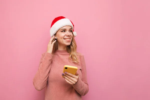 クリスマスと帽子の魅力的な幸せな女の子は、手にスマートフォンで立って、ワイヤレスヘッドフォンで音楽を聞いて、笑顔と離れてコピースペースを見ています。ピンクの背景に隔離されてるクリスマスのコンセプト. — ストック写真