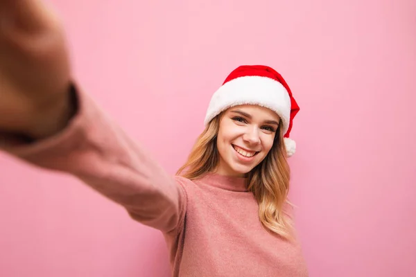 Szczęśliwa dziewczyna w świątecznym kapeluszu robi selfie na różowym tle, patrzy w aparat i uśmiecha się. Portret kobiety w kapeluszu Mikołaja robiącej zdjęcie na wyciągniętej dłoni. Koncepcja świątecznego selfie. Boże Narodzenie. — Zdjęcie stockowe