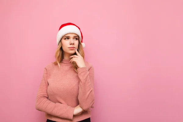 En omtänksam blondin i en julhatt står på en rosa bakgrund, tittar bort på ett tomt utrymme och tänker. Söt tjej i tomteluva tittar på kopieringsutrymme och undrade. Isolerad på rosa. Julafton — Stockfoto