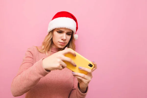 Narrated attractive gamer girl in santa claus christmas hat plays games on smartphone, looks intently at screen. Isolated on pink background. Lady plays mobile games for Christmas. X-mas — Stock Photo, Image