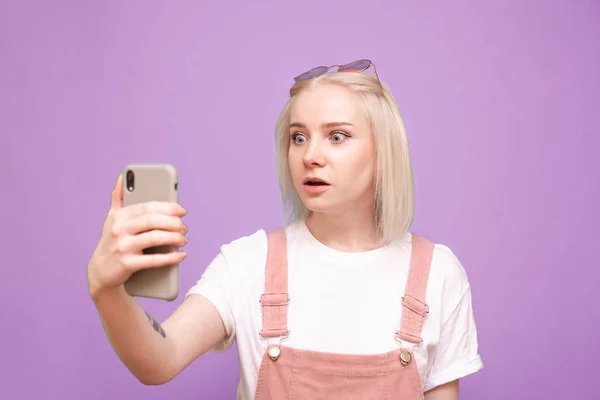 Retrato de uma menina bonito surpreso com um smartphone na mão, com o rosto espantado olhando para a tela de um telefone em um fundo roxo. Menina expressiva usa um smartphone, emoção surpresa — Fotografia de Stock