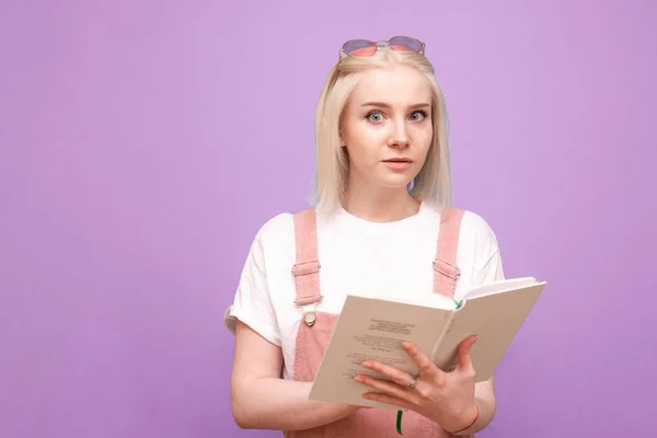 Συναισθηματικό κορίτσι με ένα βιβλίο των χεριών στέκεται σε ένα μωβ φόντο, κοιτάζοντας μέσα στην κάμερα με έκπληξη πρόσωπο.Χαριτωμένο έφηβος κορίτσι κρατά ένα βιβλίο στα χέρια της, φοράει γυαλιά, ένα λευκό T-shirt και ένα φόρεμα — Φωτογραφία Αρχείου