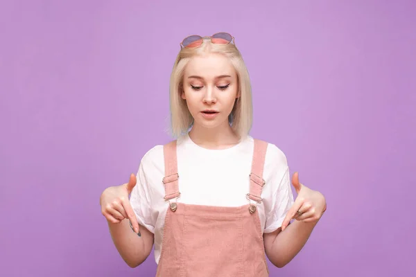 Menina loira bonita em roupas bonitos mostra os dedos para baixo e olha para um espaço vazio, isolado em um fundo roxo. bonito teen menina mostra um dedo no copyspace isolado . — Fotografia de Stock