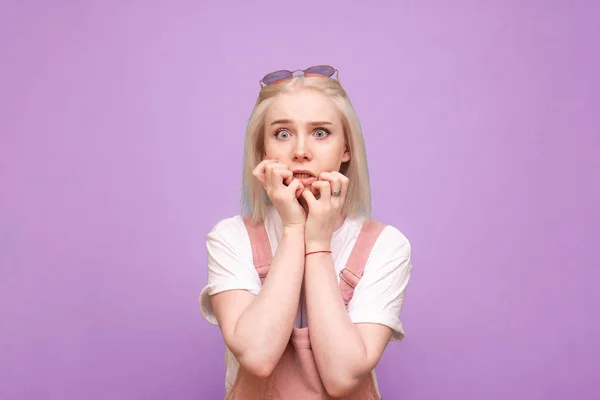 Портрет переляканої дівчини на фіолетовому тлі виглядає шокованим на камеру і кусає пальці. Ізольовані. Дівчина зі світлим волоссям у милому повсякденному одязі з шокуюче обличчя . — стокове фото