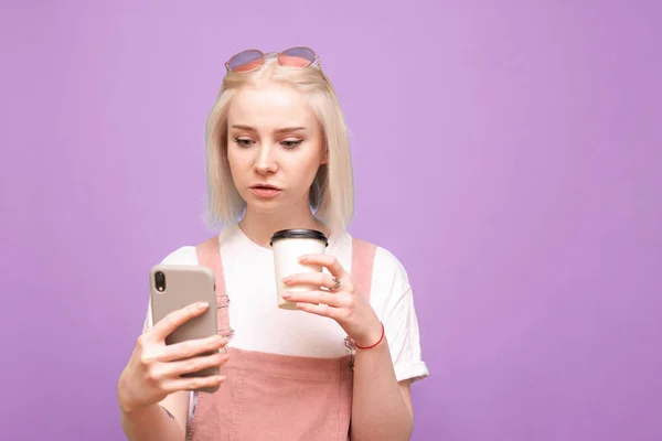 Retrato de una linda chica con una taza de café en sus manos, utilice el teléfono inteligente sobre un fondo púrpura. estudiante chica en lindo casual ropa de pie con un teléfono y una taza de café en sus manos, aislado — Foto de Stock