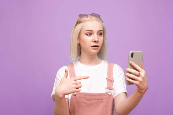 Retrato de una chica linda sorprendida muestra el dedo en el teléfono inteligente en su mano y mira la pantalla.Chica adolescente emocional en ropa linda utiliza un teléfono inteligente con una cara sorprendida sobre un fondo púrpura — Foto de Stock