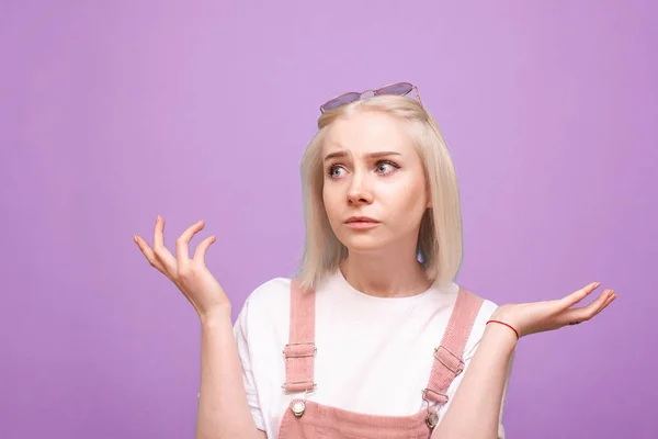 Ritratto di una ragazza perplessa bionda in abiti carini su uno sfondo viola, guardando lateralmente e mani in alto. Ritratto di donna confusa, isolata su sfondo blu . — Foto Stock