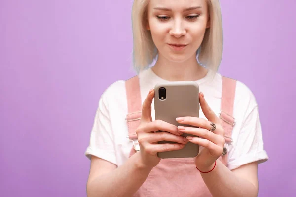 Smartphone en manos de una adolescente. Foto de cerca de una chica rubia en ropa linda utiliza un teléfono inteligente sobre un fondo púrpura, el enfoque en el teléfono en las manos de la chica. Espacio de copia — Foto de Stock