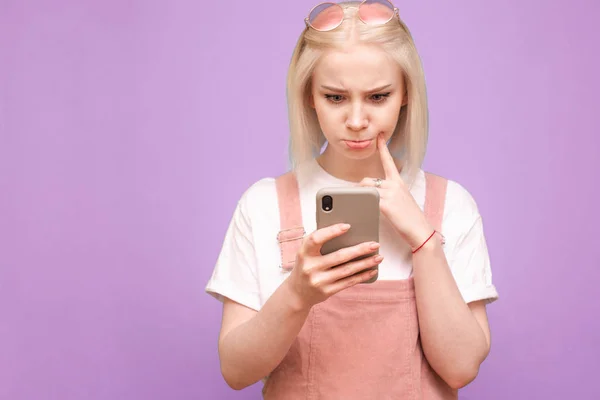 Chica rubia divertida en ropa linda con una cara reflexiva utiliza un teléfono inteligente sobre un fondo púrpura, el enfoque en el teléfono en las manos de la chica. Espacio de copia — Foto de Stock