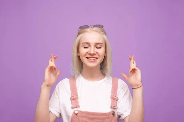 Gelukkig schattig tienermeisje doet een wens op een paarse achtergrond met haar ogen dicht, glimlacht en draagt een heldere casual kleding. geïsoleerd. — Stockfoto