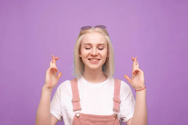 Felice ragazza adolescente carino esprime un desiderio su uno sfondo viola con gli occhi chiusi, sorride e indossa un abbigliamento casual luminoso. Isolato . — Foto Stock